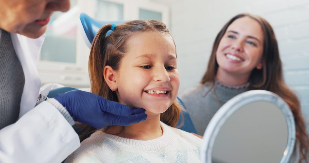 Young girl smiling at her teeth in a mirror during a dental visit with her mother nearby.